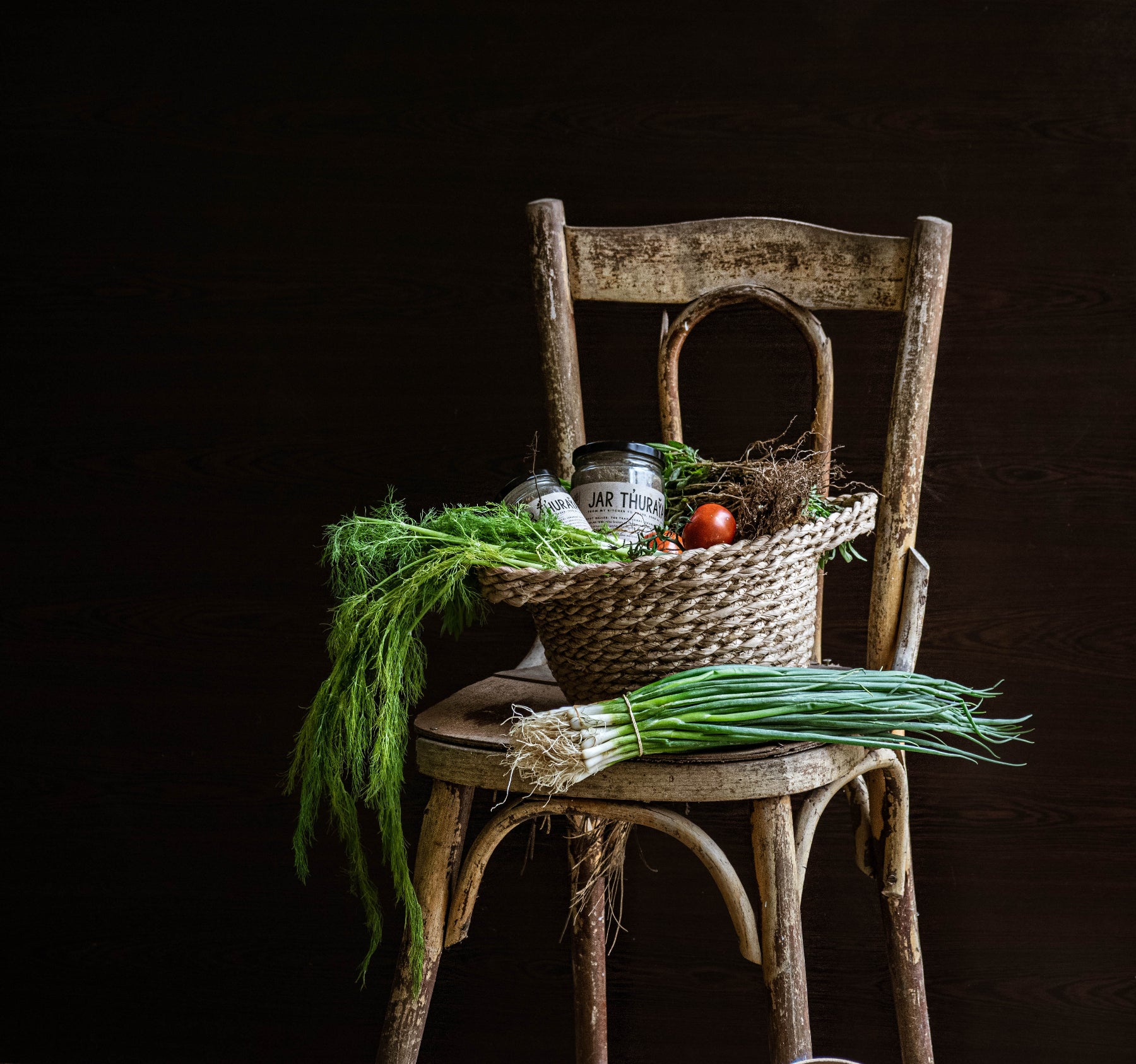 Wicker basket of fresh herbs, spring onions and a jar of Jar Thuraya za'atar on a rustic wooden chair