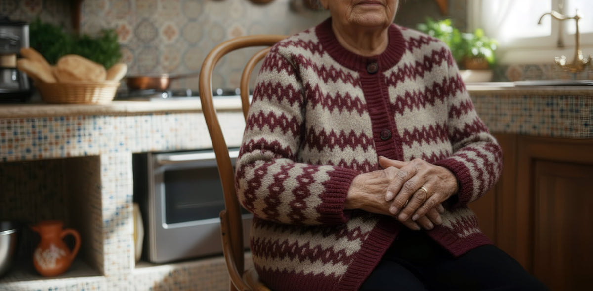 Older Lebanese woman in a burgundy knit cardigan sitting with folded hands in a traditional tiled kitchen