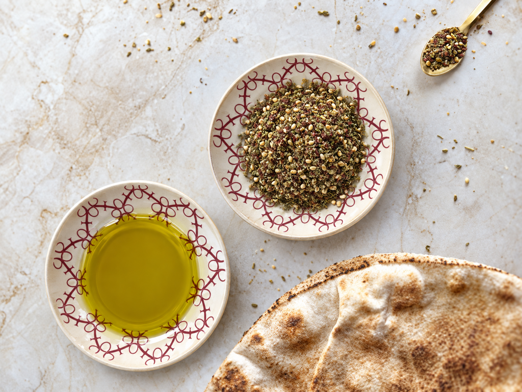 Two small patterned ceramic bowls on a light stone surface, one filled with golden olive oil and the other with coarse ground seeds or spices. A partially visible flatbread and a small spoon of the spice sit nearby.